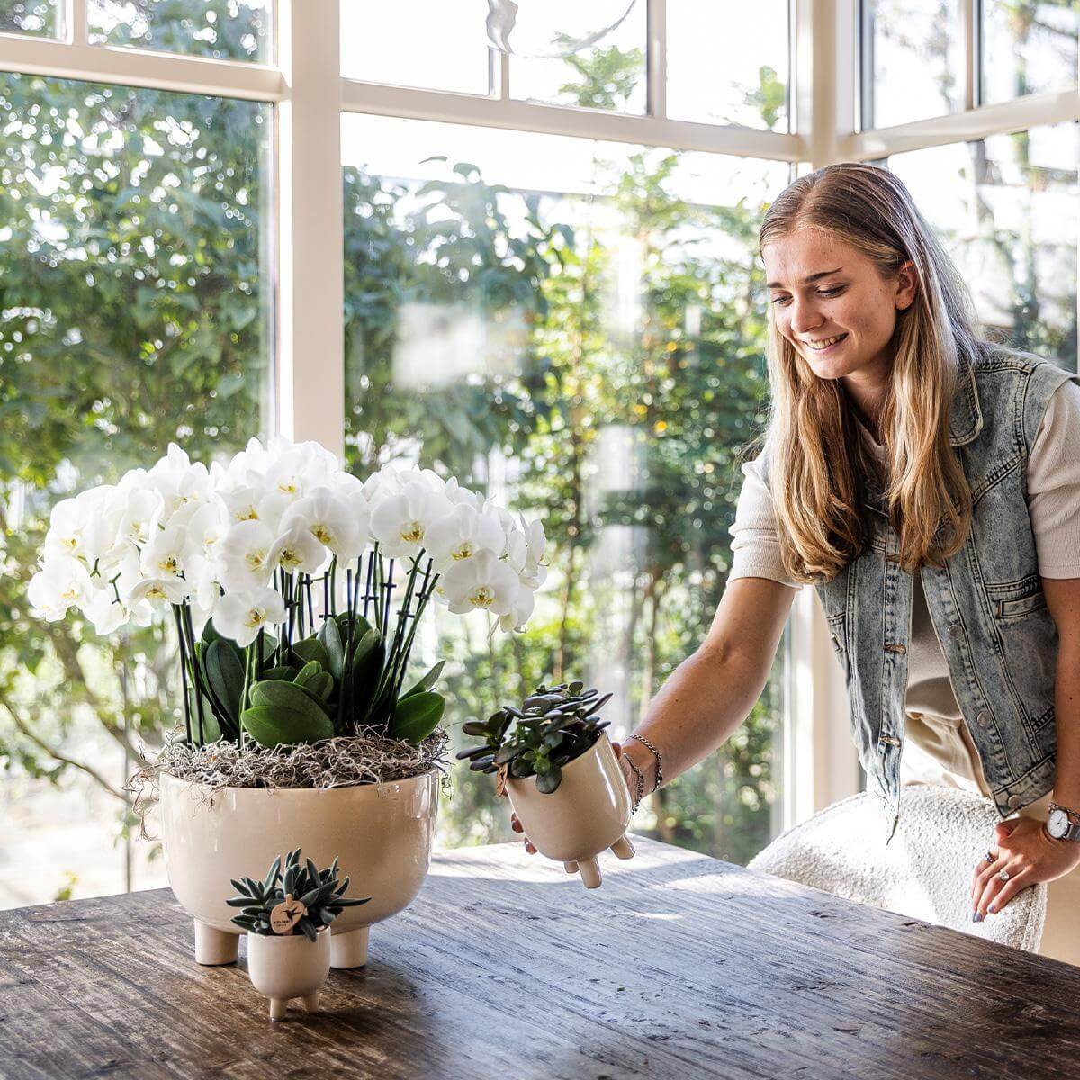 Vrouw verzorgt witte orchideeënset in keramieken schaal met waterreservoir en natuurlijke uitstraling op houten tafel.