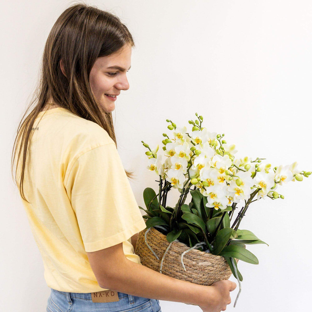 Vrouw houdt een Kolibri Orchids witte plantenset in Reed Basket met waterreservoir vast, inclusief drie witte orchideeën Lausanne en drie groene planten.