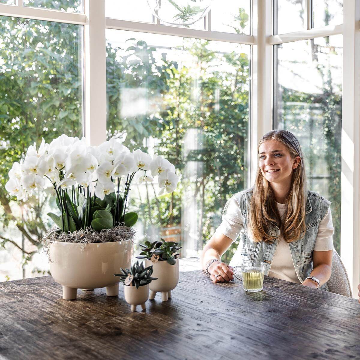 Vrouw geniet van een kopje thee naast een schaal met witte orchideeën in een zonnige kamer.