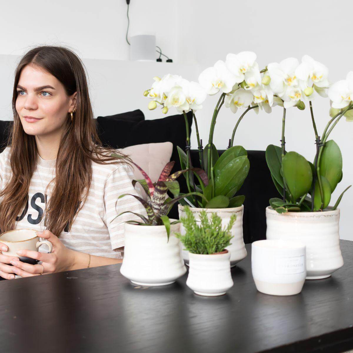 Vrouw met kop koffie naast witte Phalaenopsis orchideeën in witte potten, sfeervolle huiskamerdecoratie.