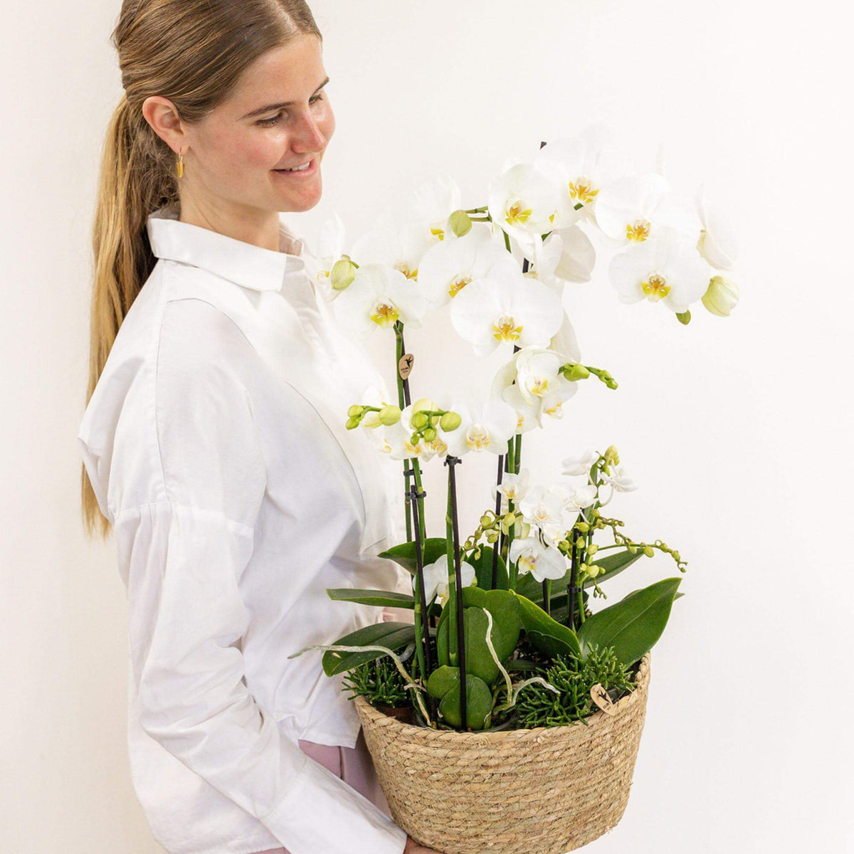 Vrouw met witte Kolibri Orchids en groene planten in rieten mand met waterreservoir, Field Bouquet wit.