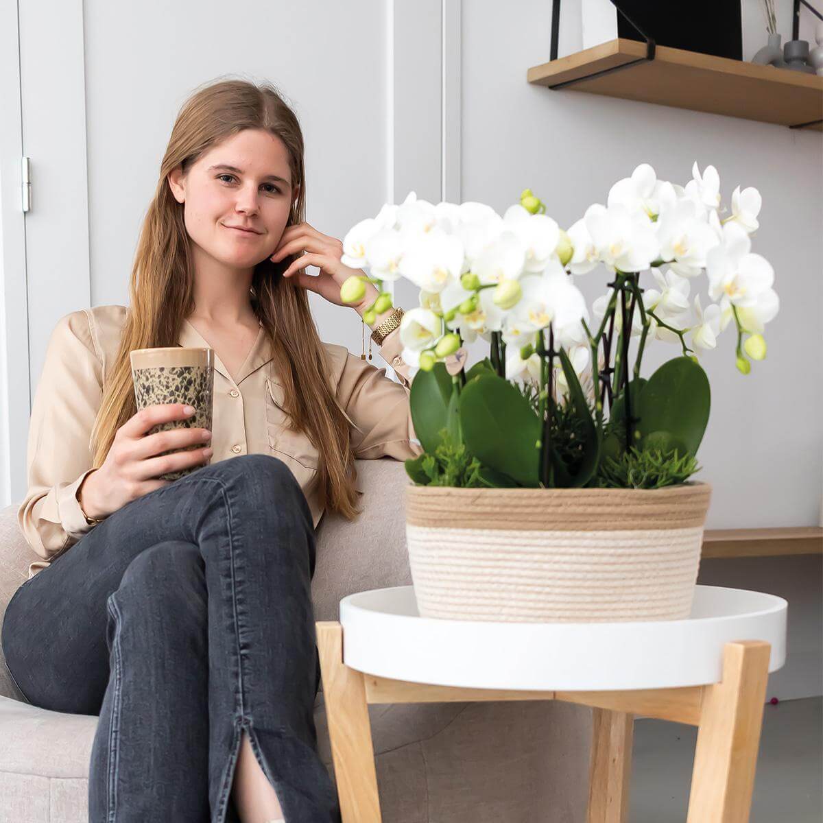 Vrouw geniet van koffie naast Kolibri Orchids set met drie witte orchideeën in katoenen mand, op tafel in moderne woonkamer.