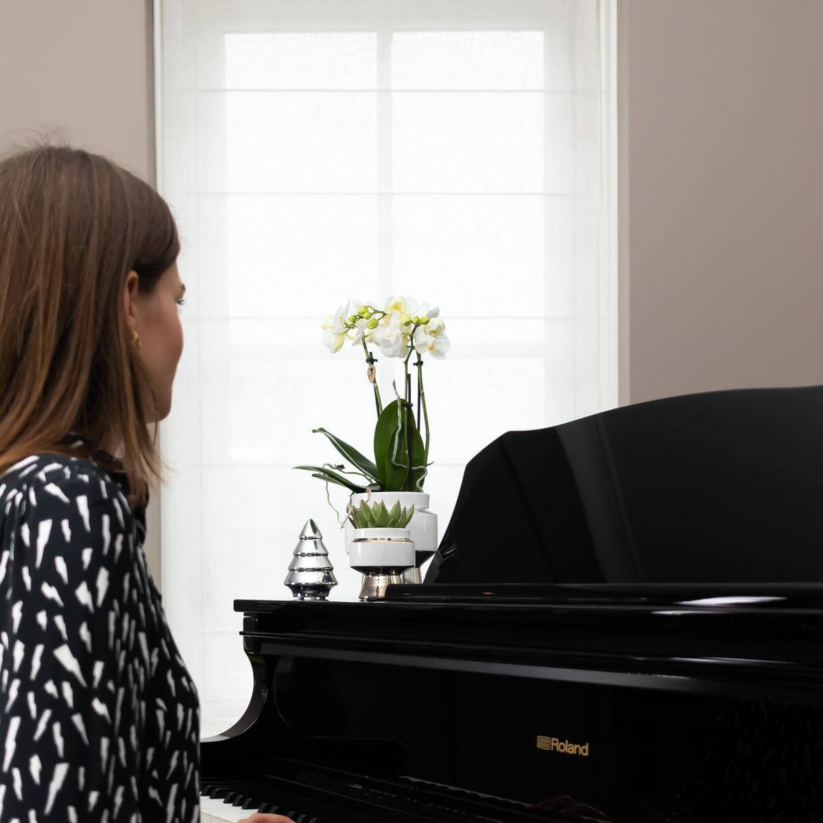 Vrouw speelt piano met witte Phalaenopsis orchidee in zilveren Le Chic sierpot op de achtergrond