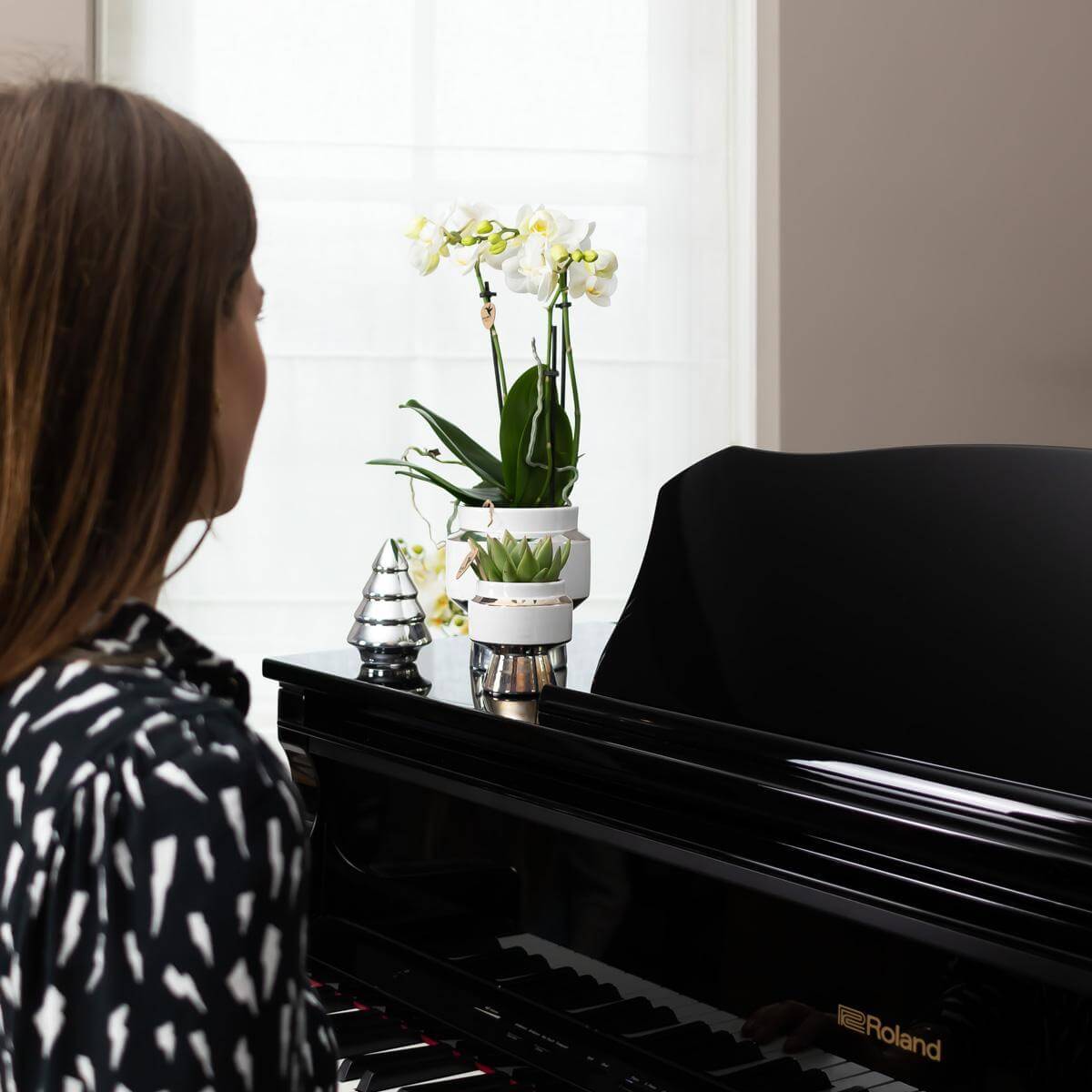 Vrouw geniet van Witte Phalaenopsis orchidee in zilveren Le Chic sierpot naast zwarte piano.